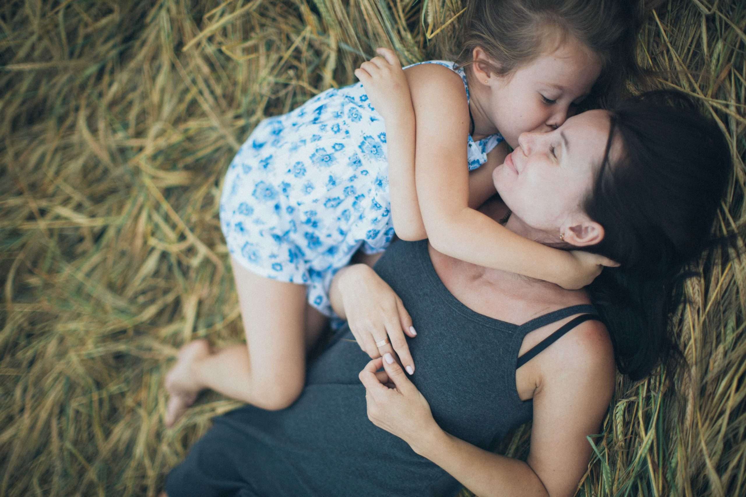 Niña besando a su madre, tumbadas en un campo verde, felices y tranquilas. Educar Positivo - Maternidad Feliz- by Elina-sazonova